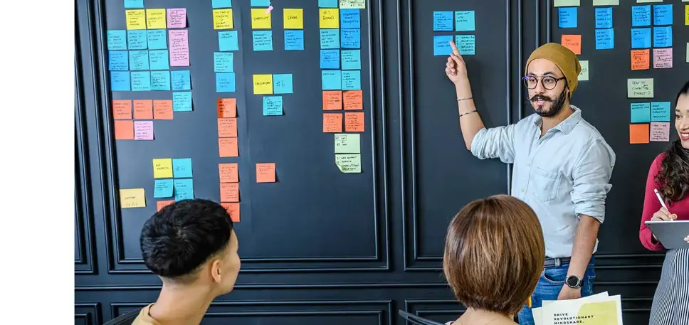 Three seated people are looking at a wall covered with post it notes, set out in columns, while a man stands and points to the notes explaining